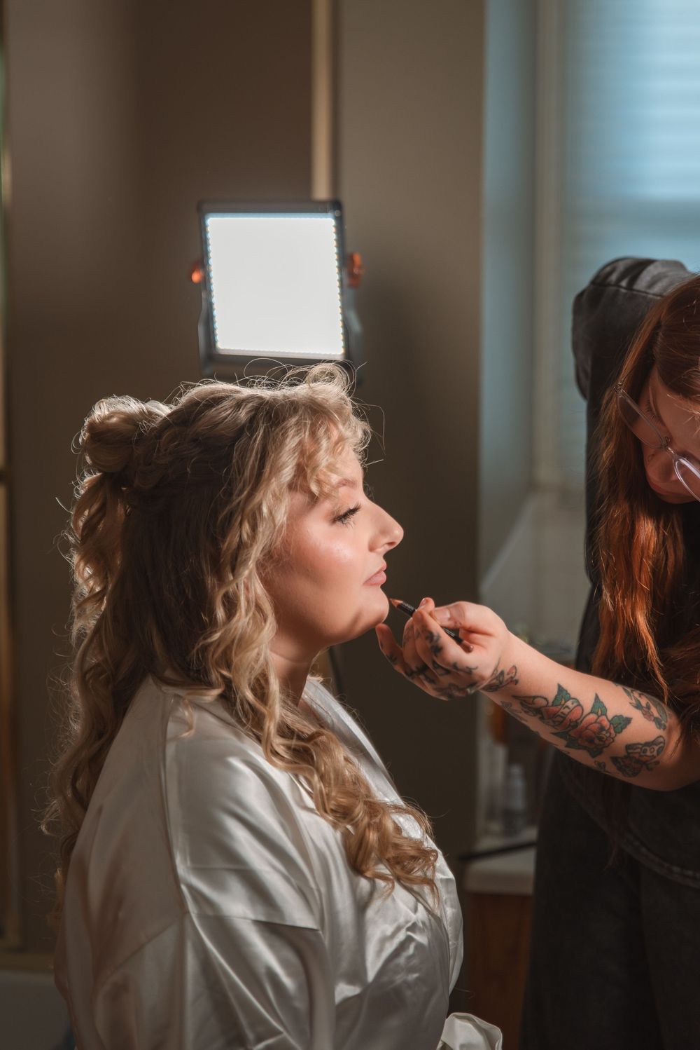 A makeup artist with tattoos applies lipstick to a bride with curly hair under a bright ring light.