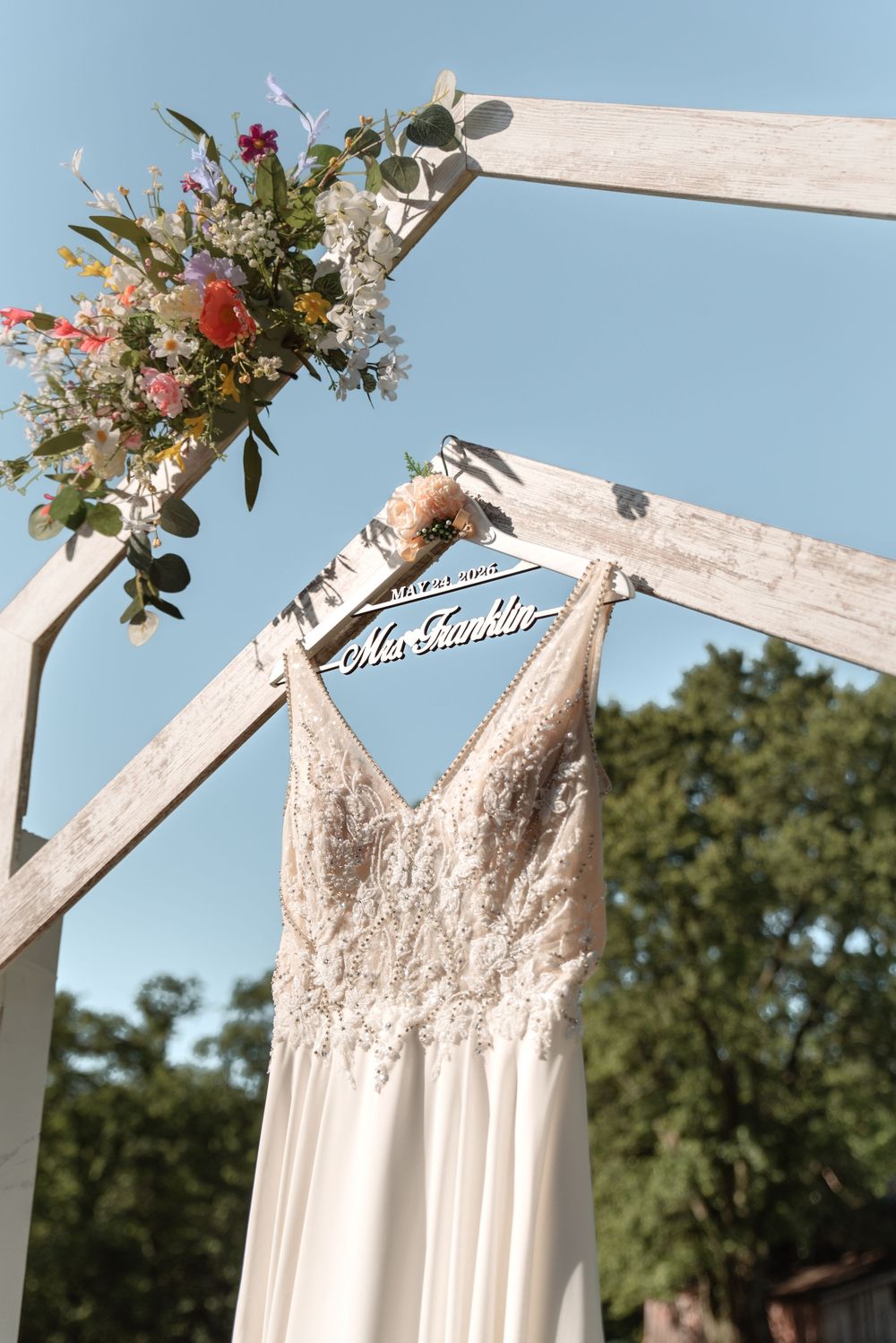 Elegant lace wedding dress hanging on a personalized Mrs. hanger from a rustic white wooden arch with floral decor.