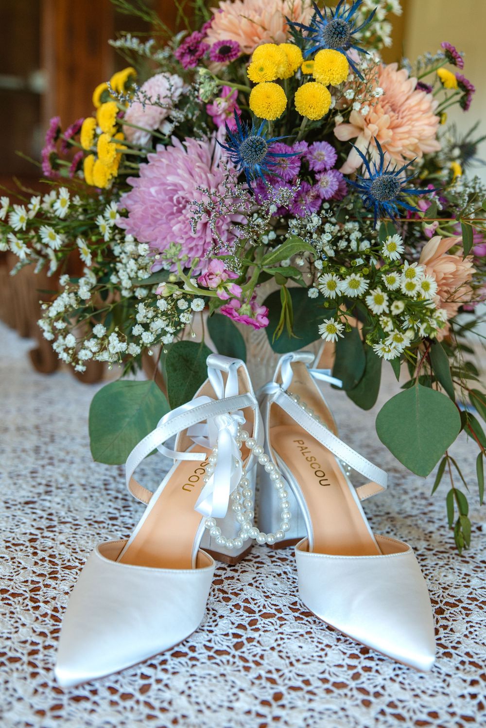 White satin bridal heels with ribbon ties displayed on lace beside a colorful wedding bouquet of wildflowers.