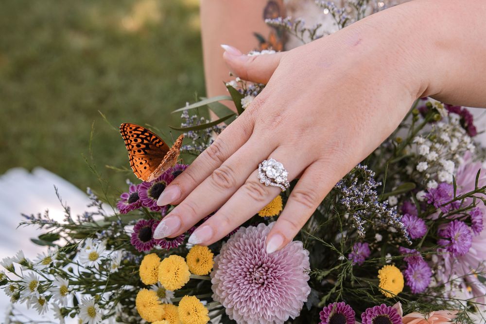 Close-up of a diamond engagement ring on a hand resting on a colorful bouquet with an orange butterfly.