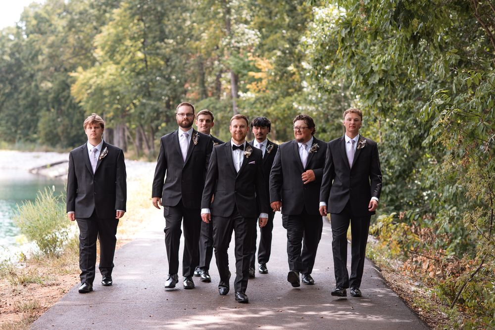 Groom and groomsmen in black suits walking together on a path surrounded by trees at an outdoor wedding venue.