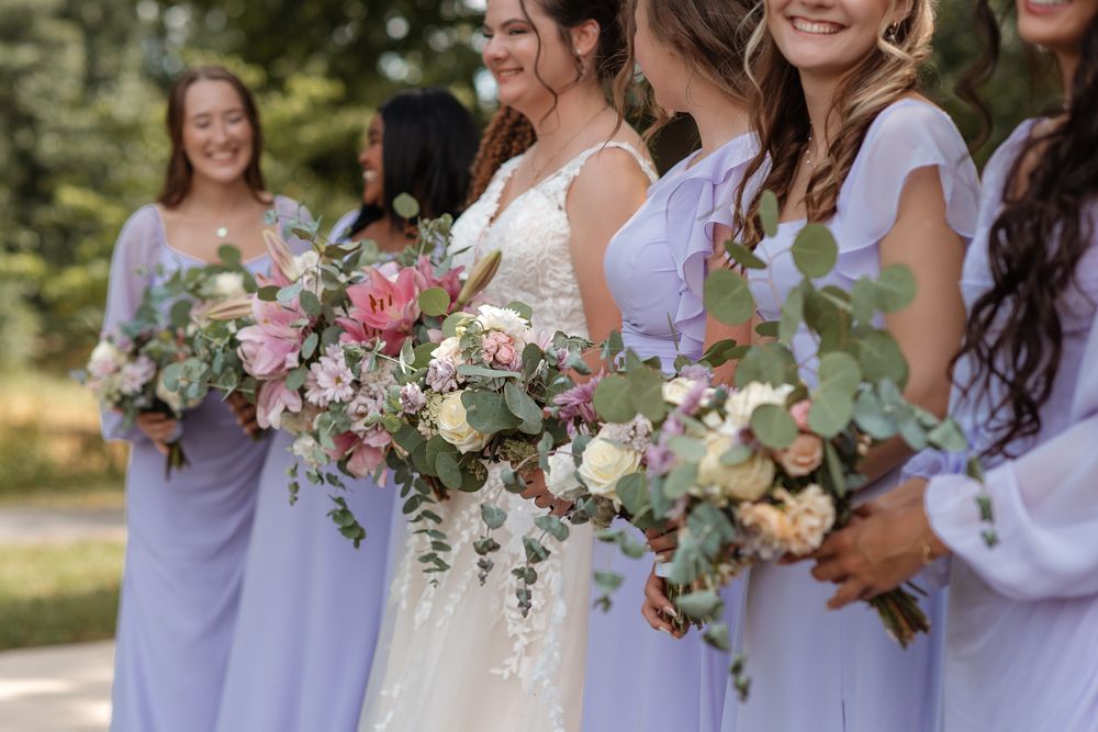Bride in white lace gown with bridesmaids in lavender dresses holding lush floral bouquets with eucalyptus outdoors.