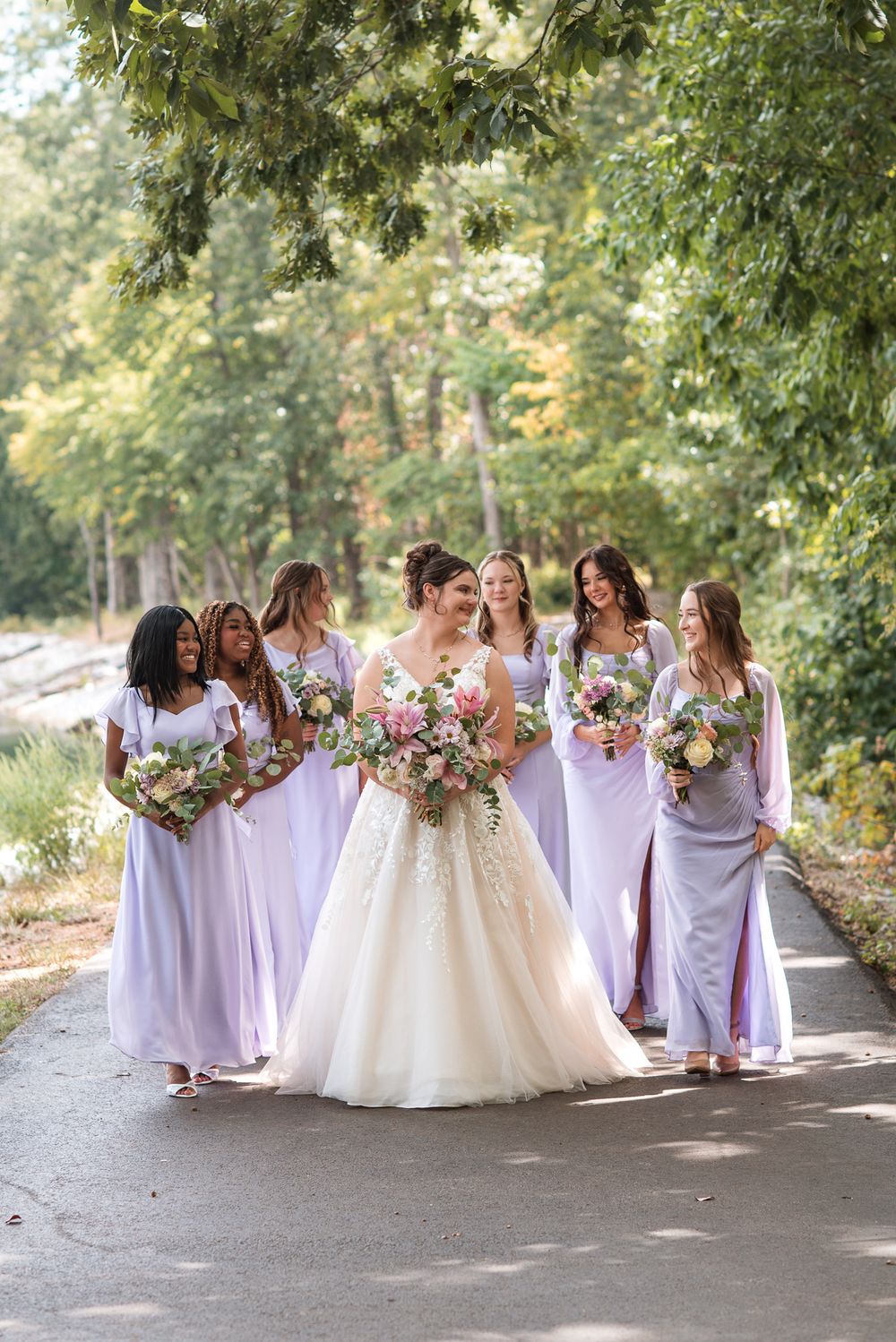 Bride in white gown walks with bridesmaids in lavender dresses, holding floral bouquets on a tree-lined path.