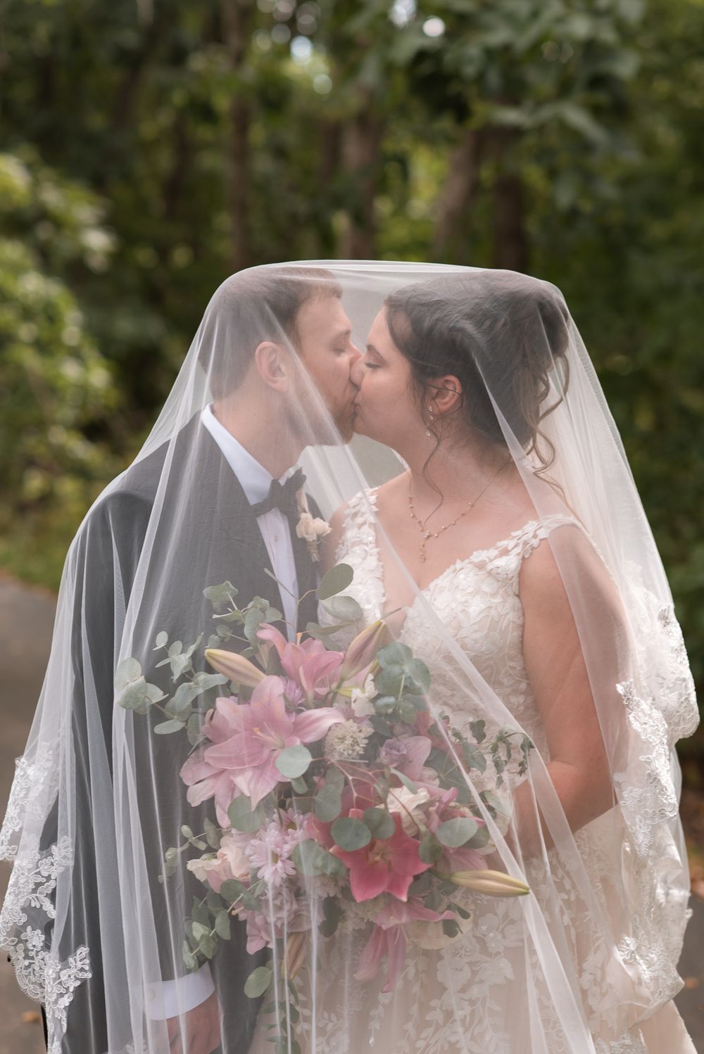Bride and groom kissing under a lace wedding veil, bride holding a pink lily and eucalyptus bouquet outdoors.