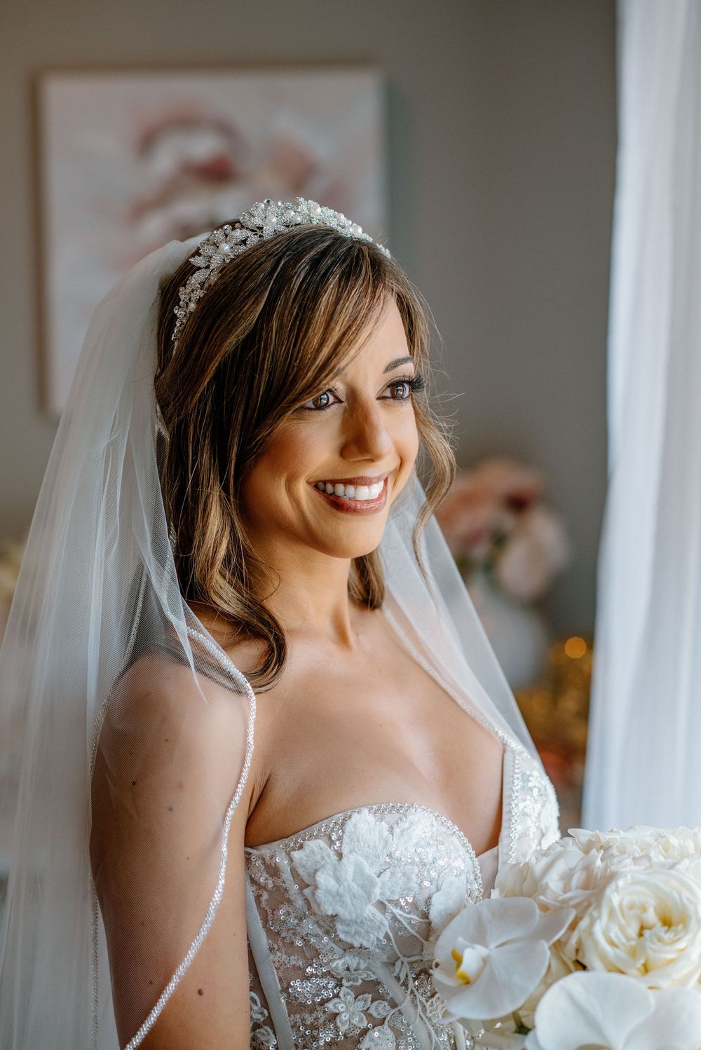 A radiant bride in a white lace gown and crystal tiara smiling by a window.