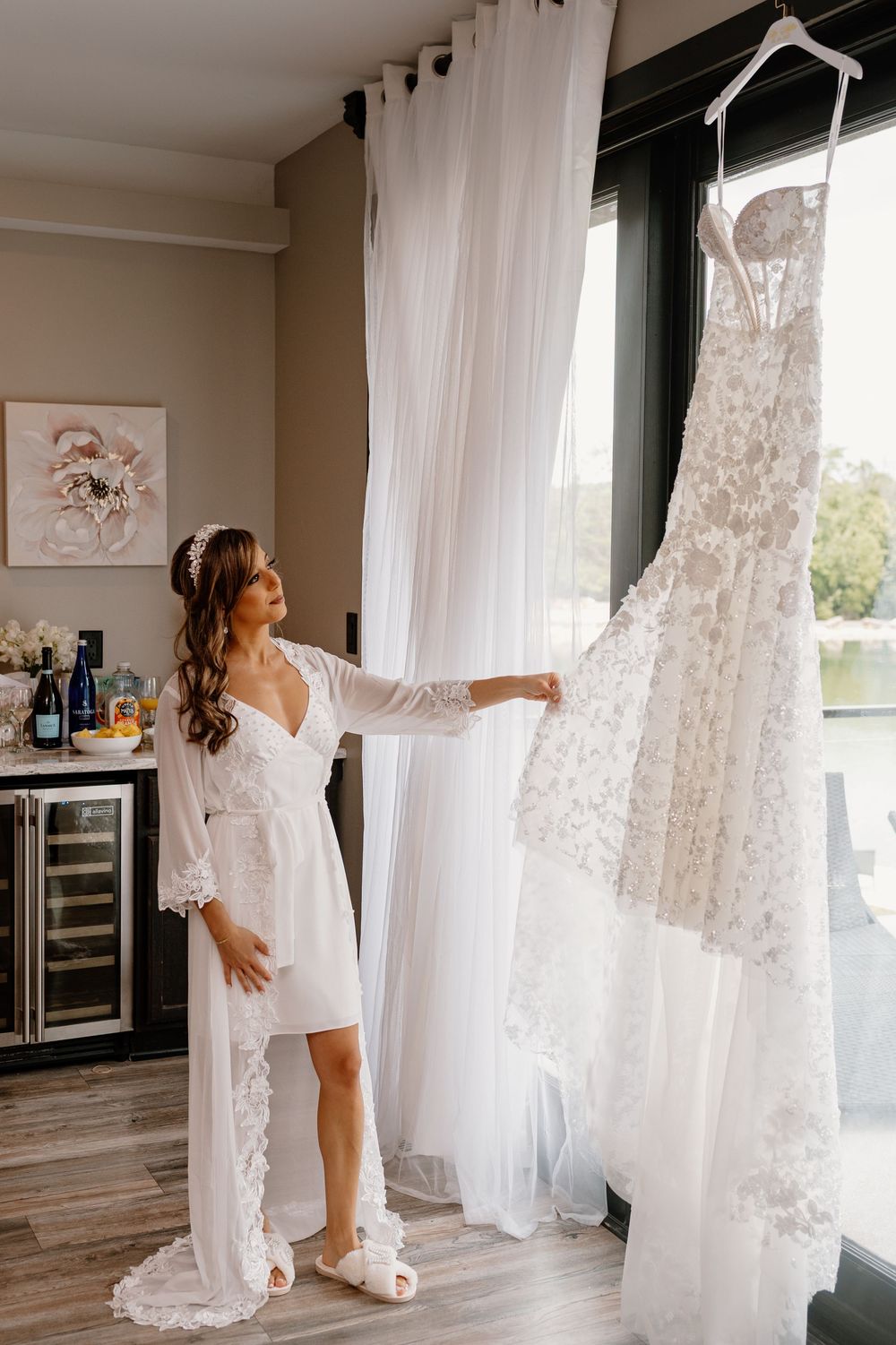 Wedding dress hanging in bridal suite with romantic morning light streaming through sheer white curtains.