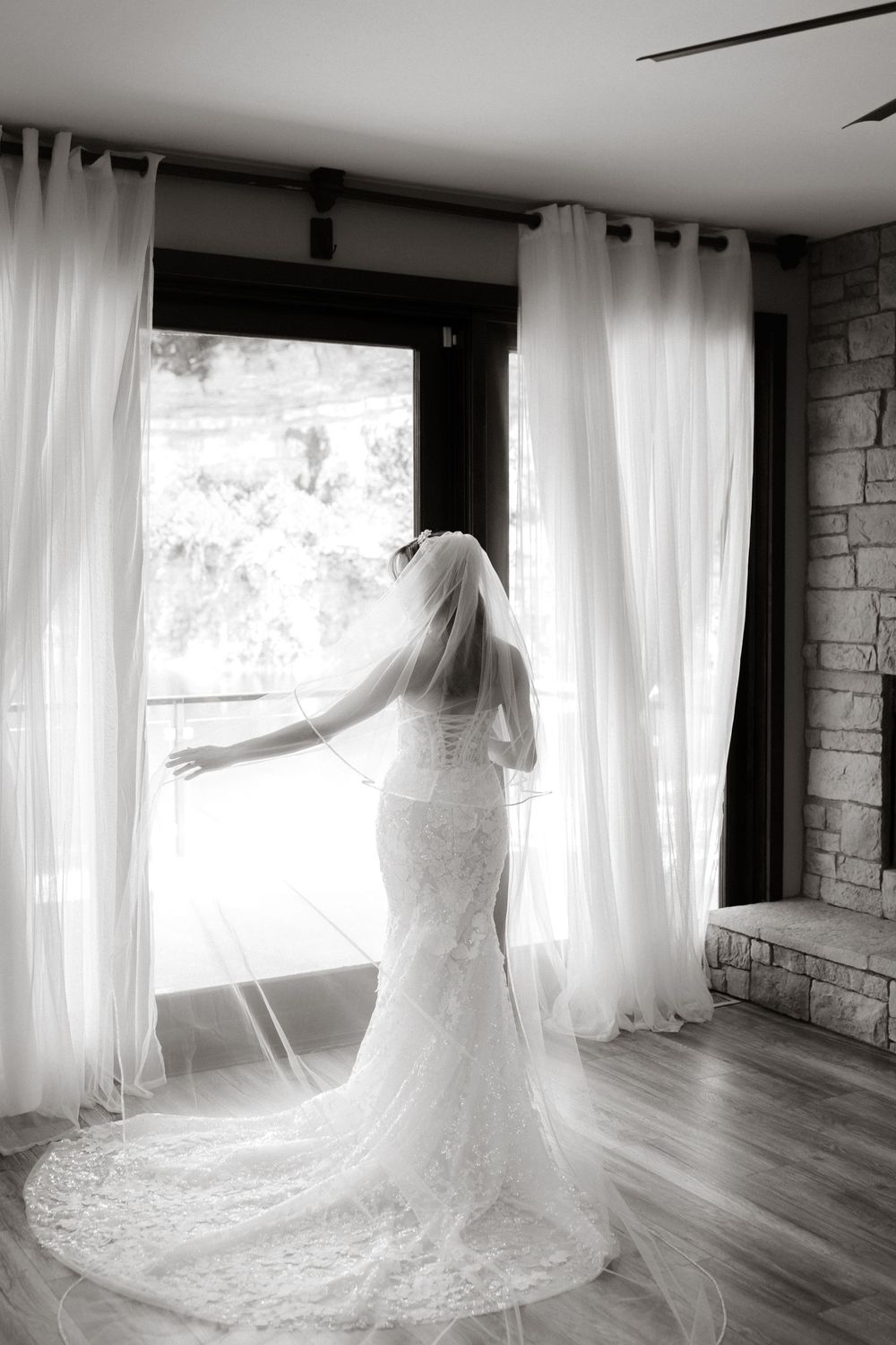 A bride in a lace wedding gown with long train stands gracefully by sheer white curtains in natural light.