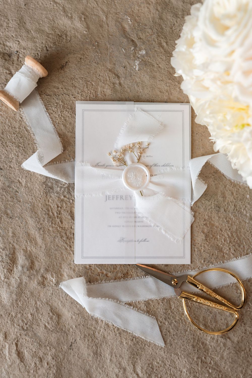 Elegant white wedding invitation suite with silk ribbon and gold scissors on textured stone surface.