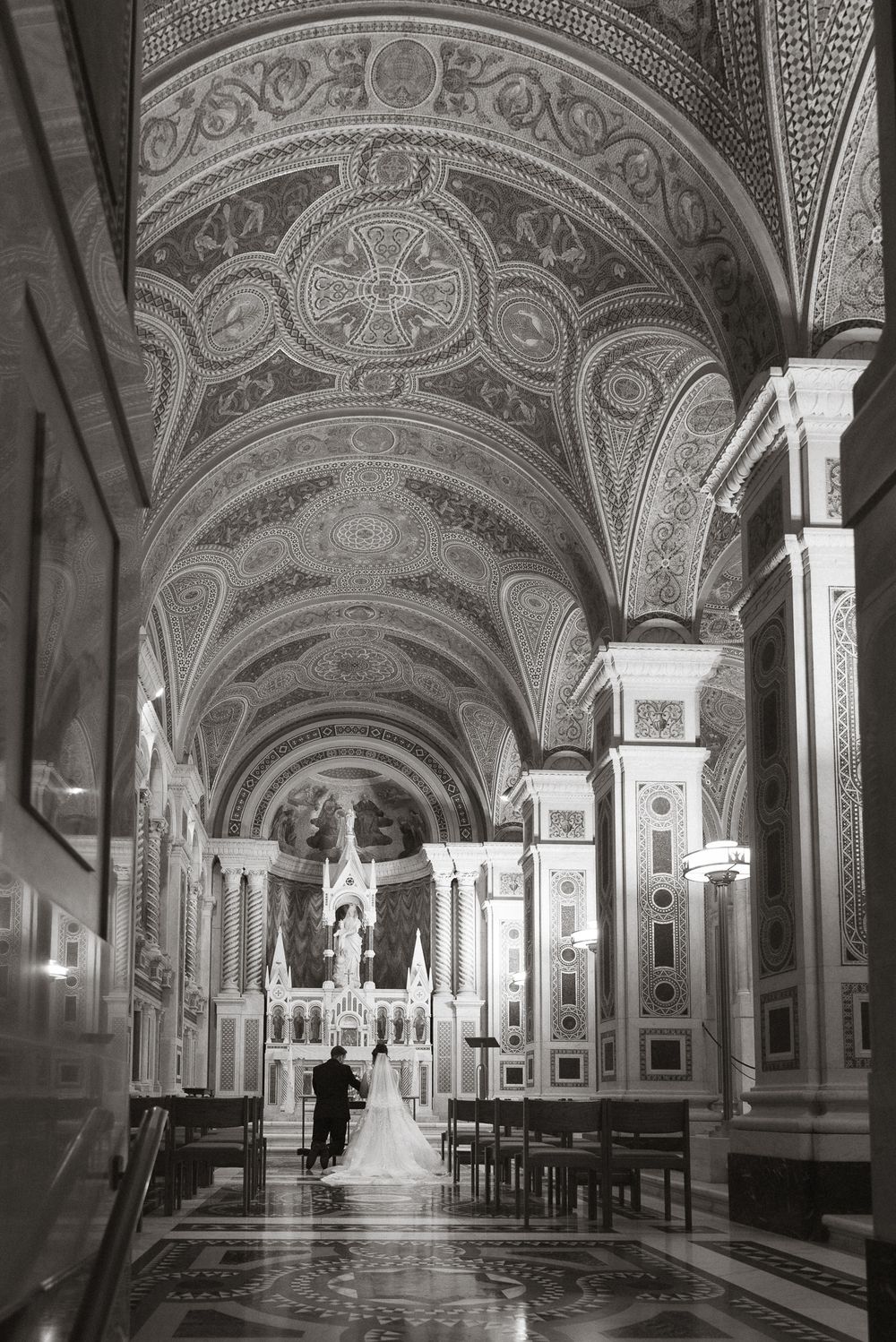 Black and white photo of a bride and groom at the altar of an ornate cathedral with vaulted ceilings.