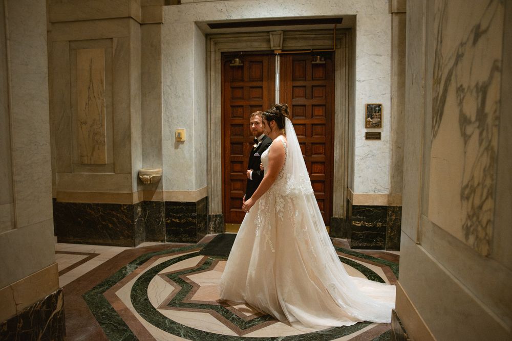 Bride in elegant white wedding gown with train stands before ornate wooden doors in a grand marble-floored venue.