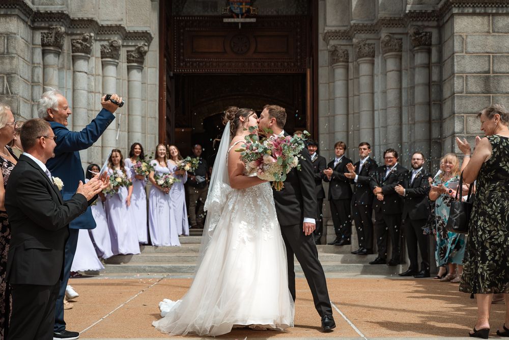 Newlywed couple sharing first kiss outside historic church, surrounded by cheering guests and bridesmaids in lavender gowns.