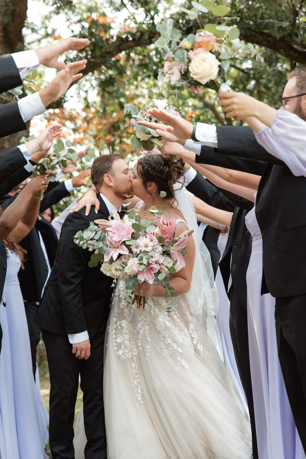 Bride and groom share a kiss while wedding party raises bouquets overhead in a romantic outdoor ceremony celebration.