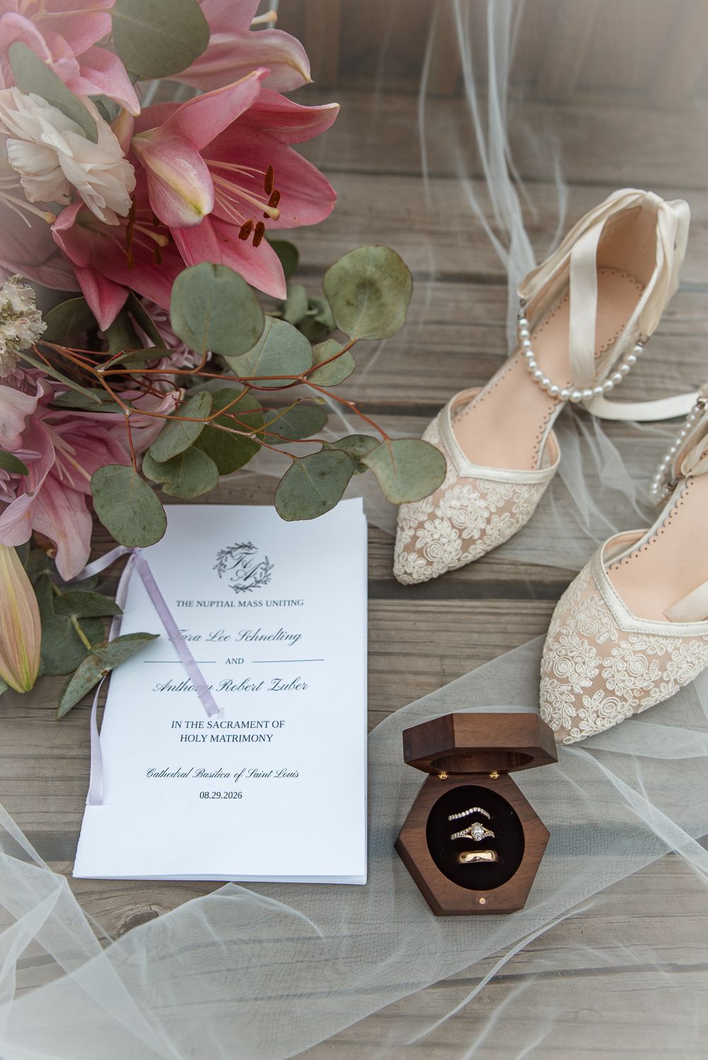 Elegant wedding flat lay with lace bridal heels, floral bouquet, ceremony program, and gold rings in a velvet box on wood.