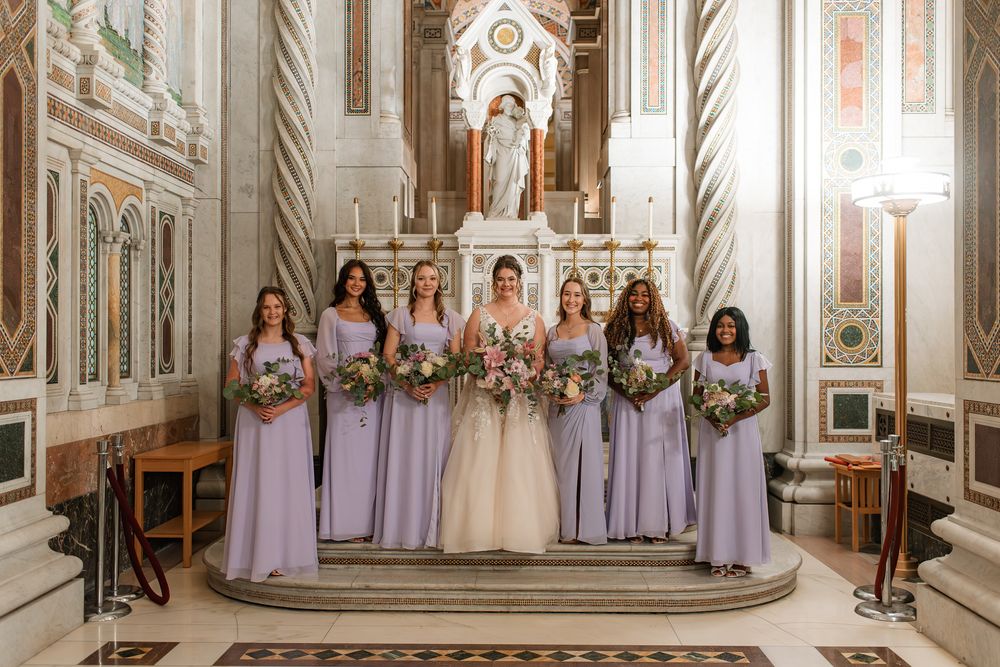 Bride and bridesmaids in lavender gowns holding bouquets inside an ornate church altar.