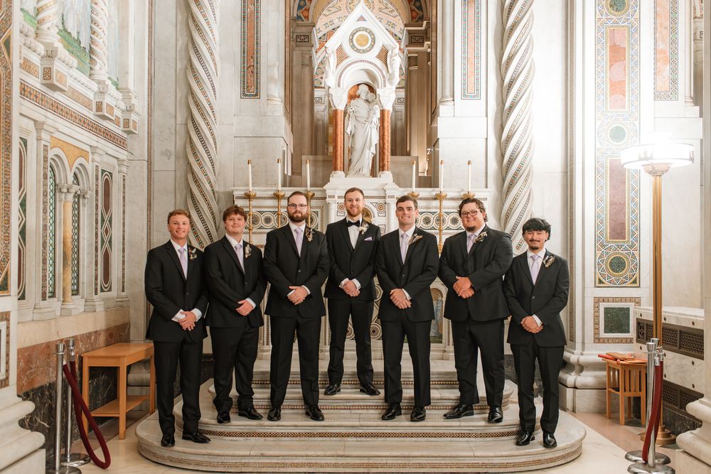 Groom and groomsmen in black suits posing together inside an ornate church with marble altar backdrop.