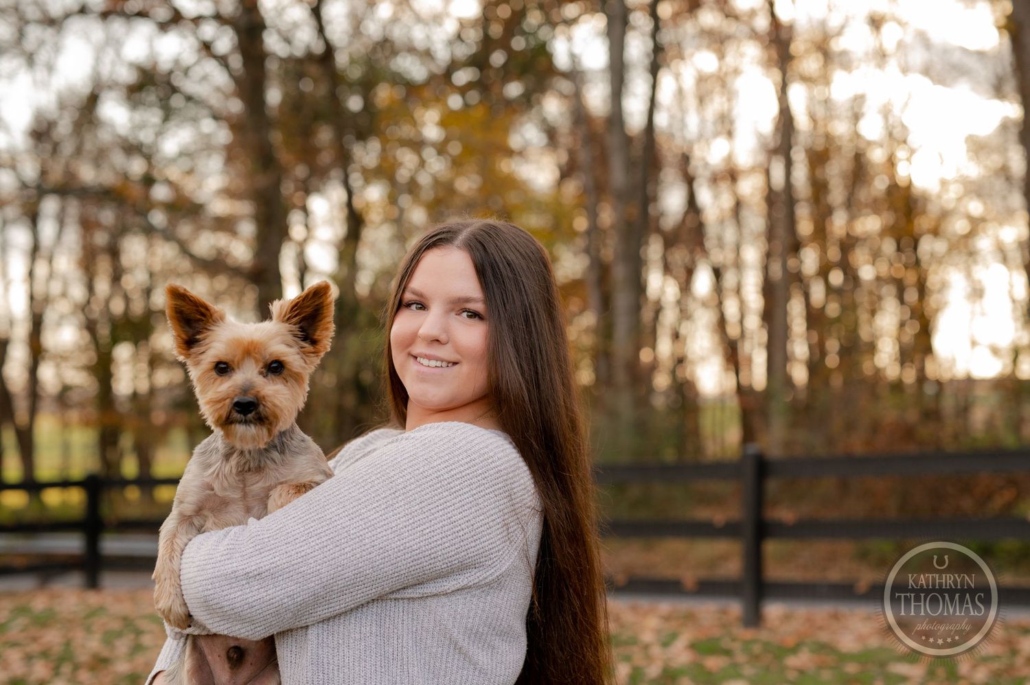 Person holding a Yorkshire Terrier dog while posing in front of autumn trees and wooden fencing.