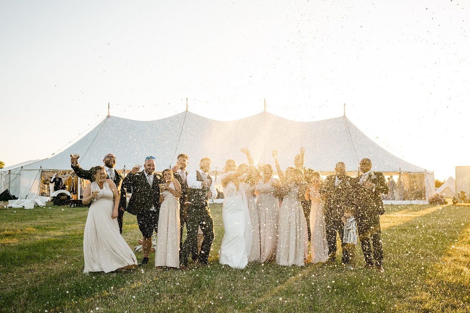 Wedding party poses together in front of a white tent during golden hour sunset at an outdoor reception wedding tent in Nova Scotia.