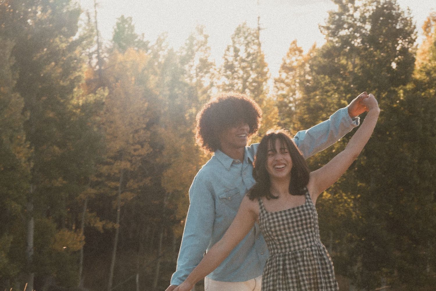 A romantic couple dances playfully in a golden sunlit forest while wearing casual clothing at sunset.