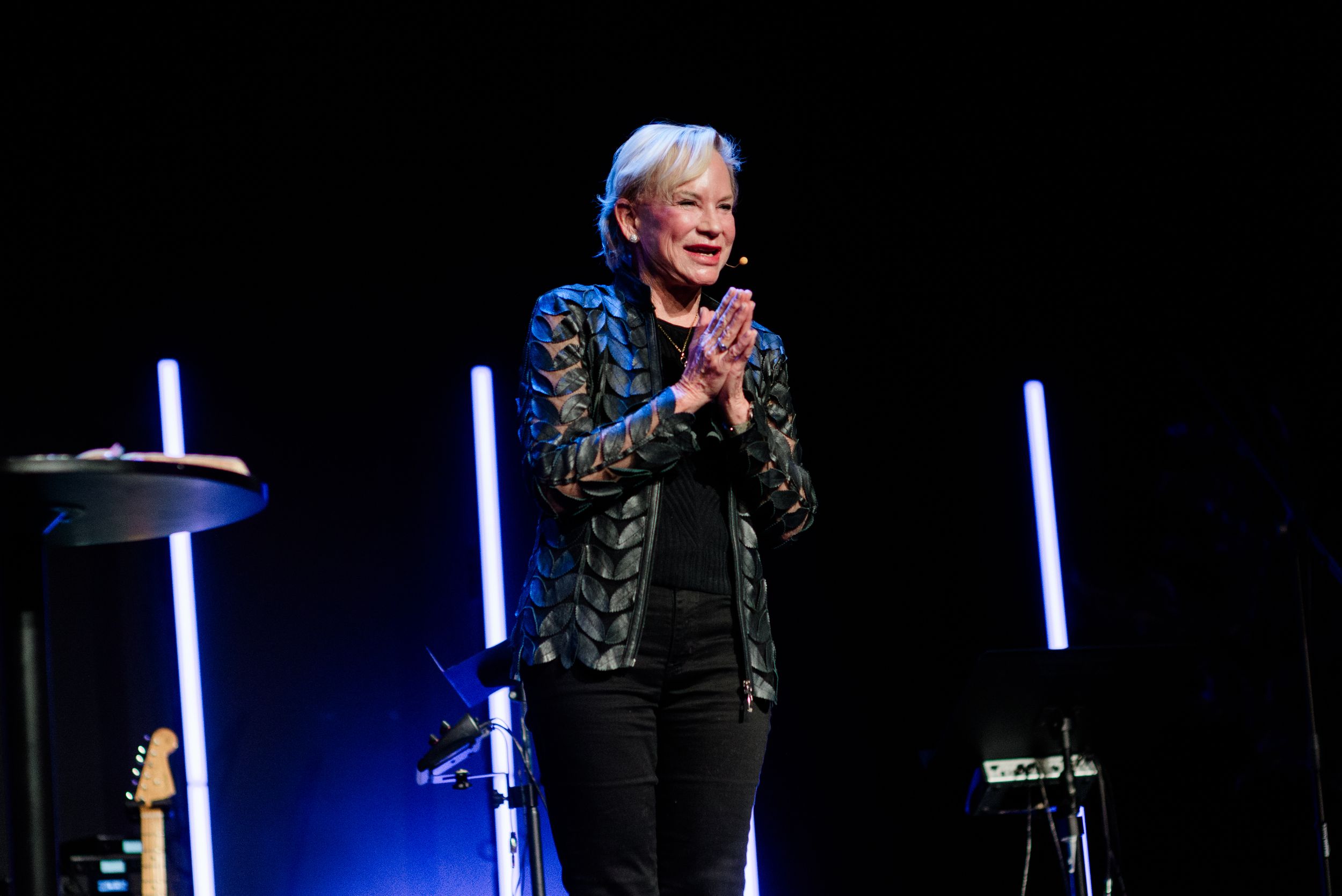 Woman in black jacket clapping on stage with neon lights at a live event.