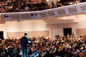 Graduate in cap and gown addresses a large audience in a packed auditorium during a graduation ceremony.