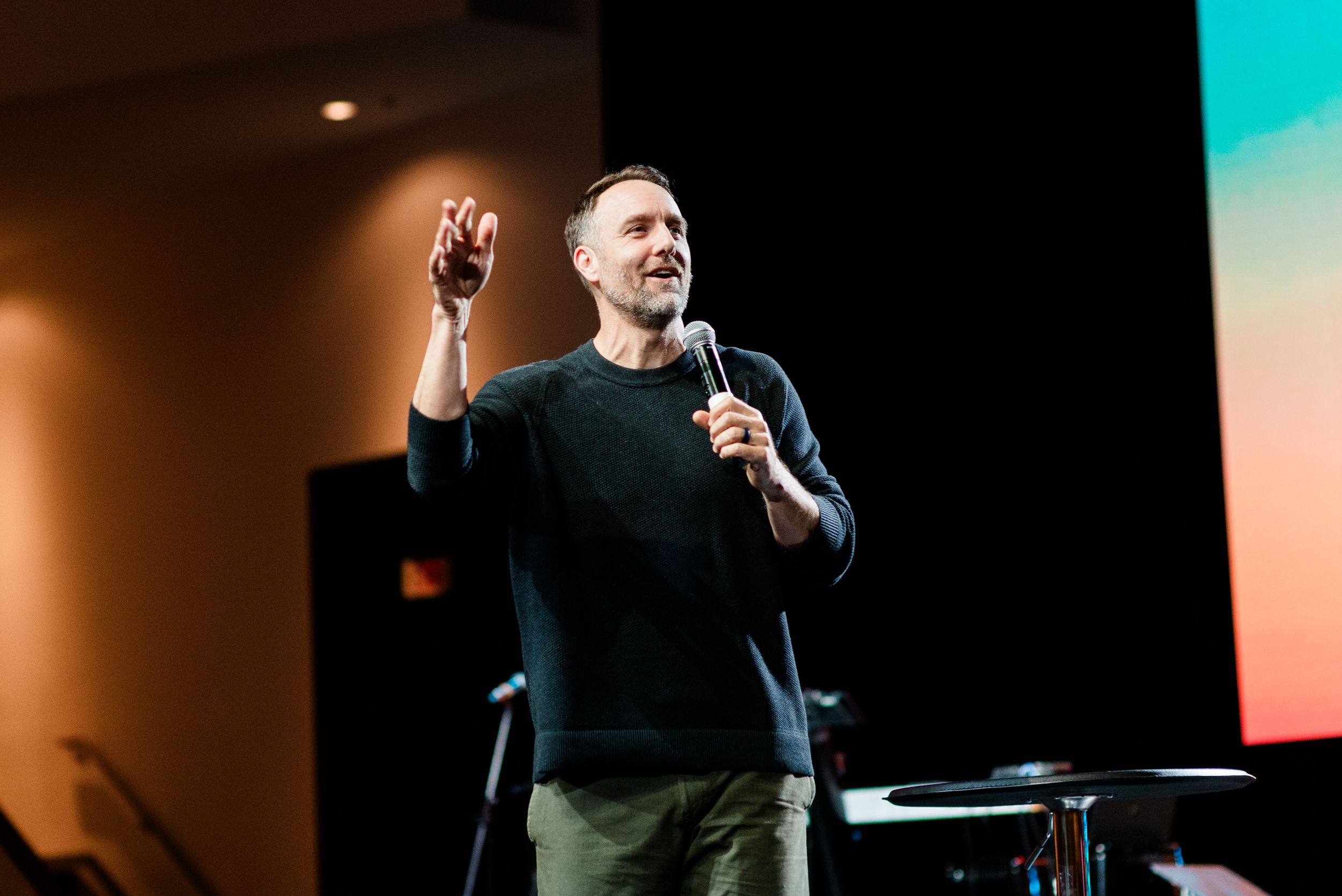 Man speaking on stage holding microphone with hand raised in modern auditorium with colorful backdrop.