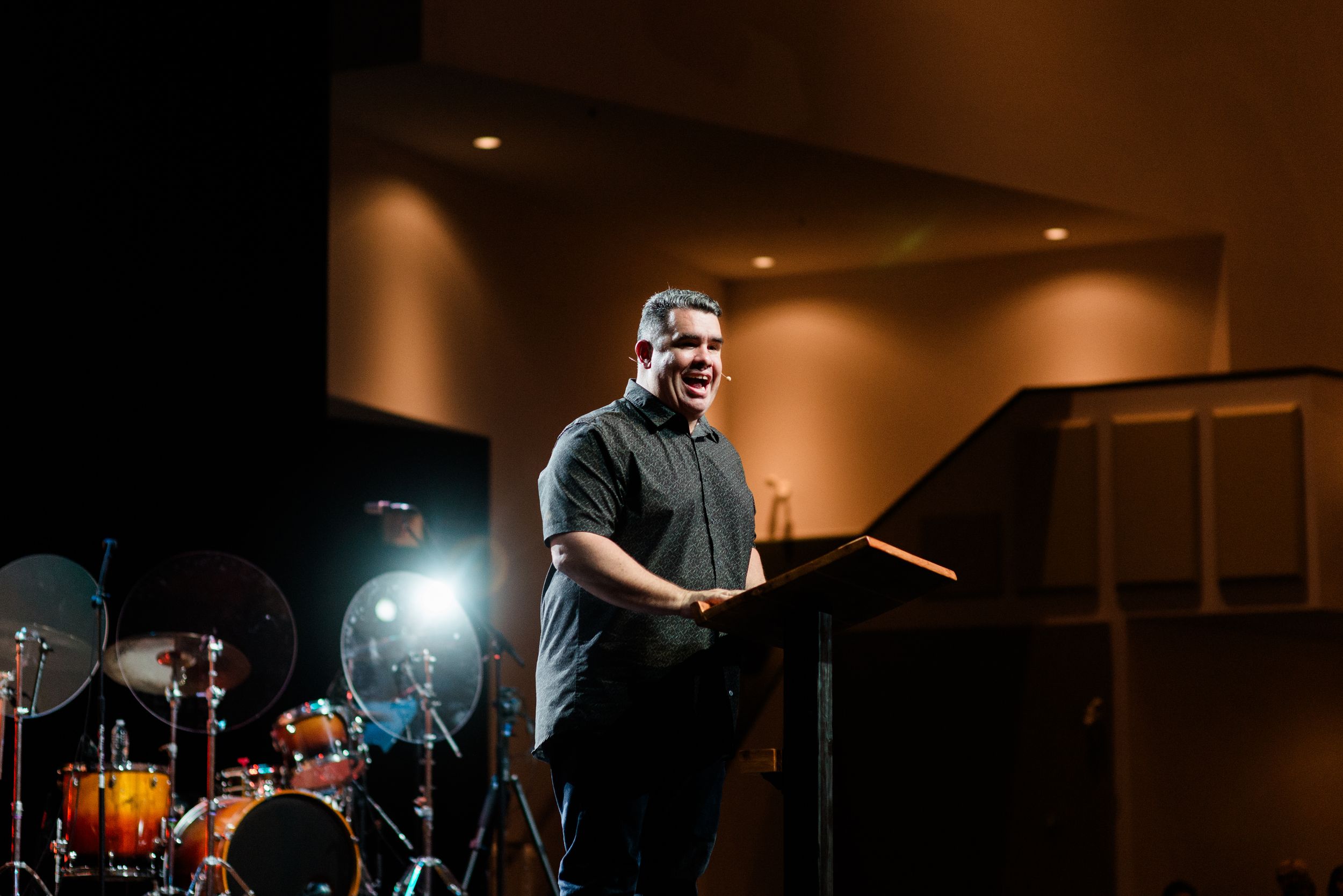 Man in black shirt speaking at podium on stage with drum kit and bright stage lighting behind him.