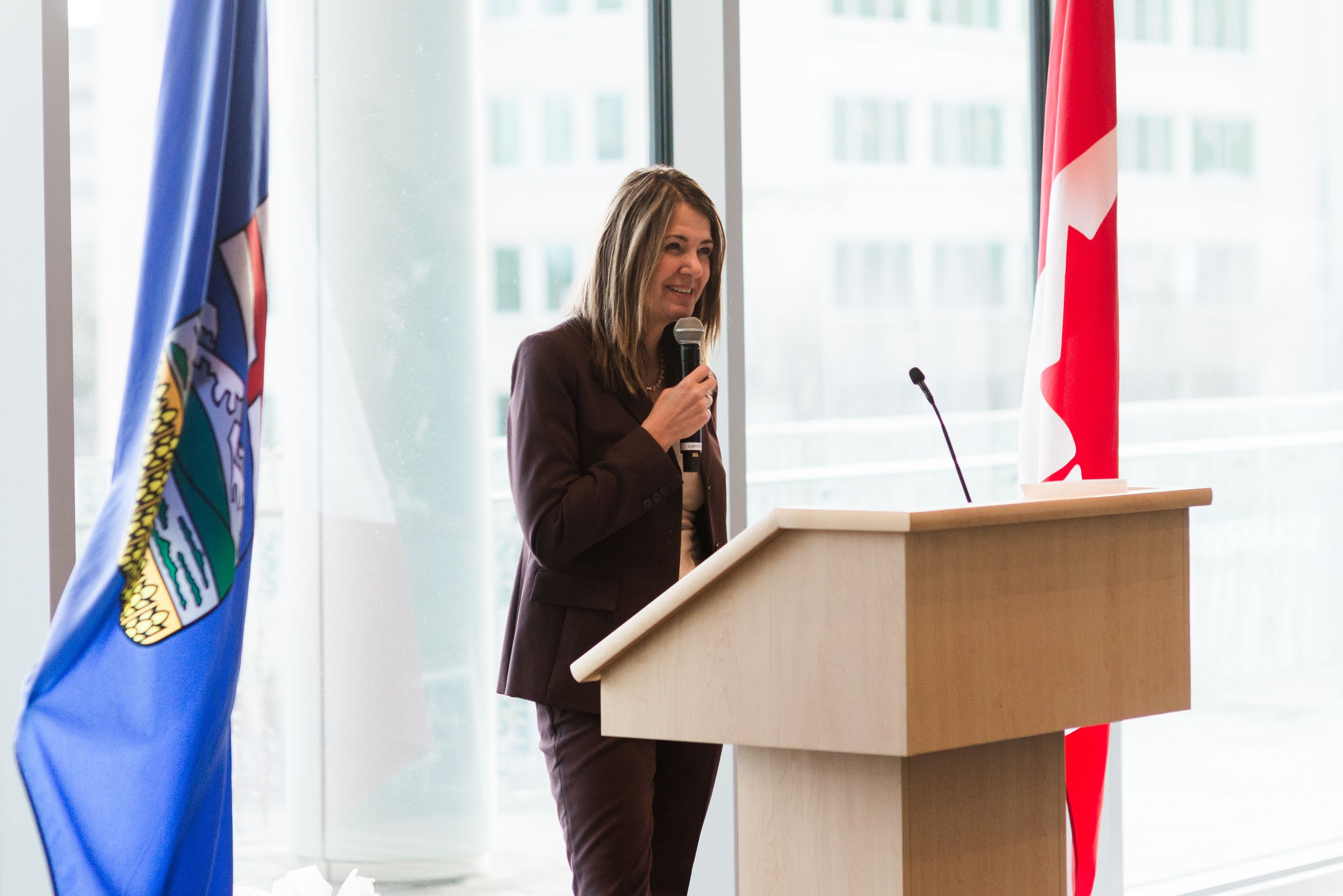 Woman in brown suit speaking at podium with Alberta and Canadian flags in modern office building.