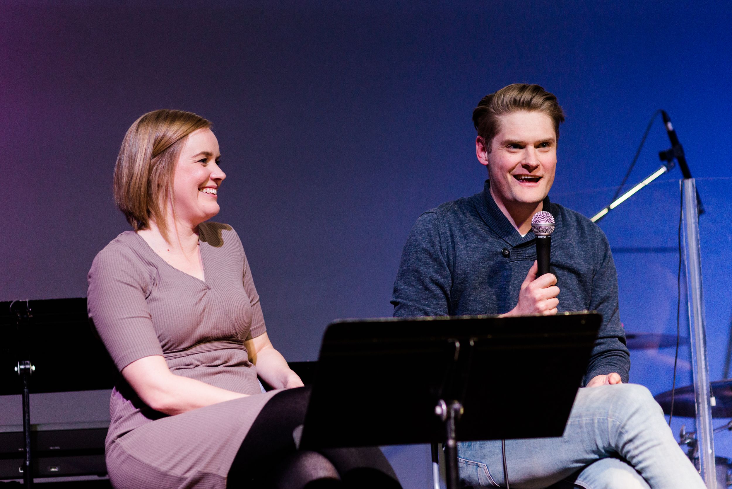 Two speakers seated at podiums on stage during presentation with blue lighting.