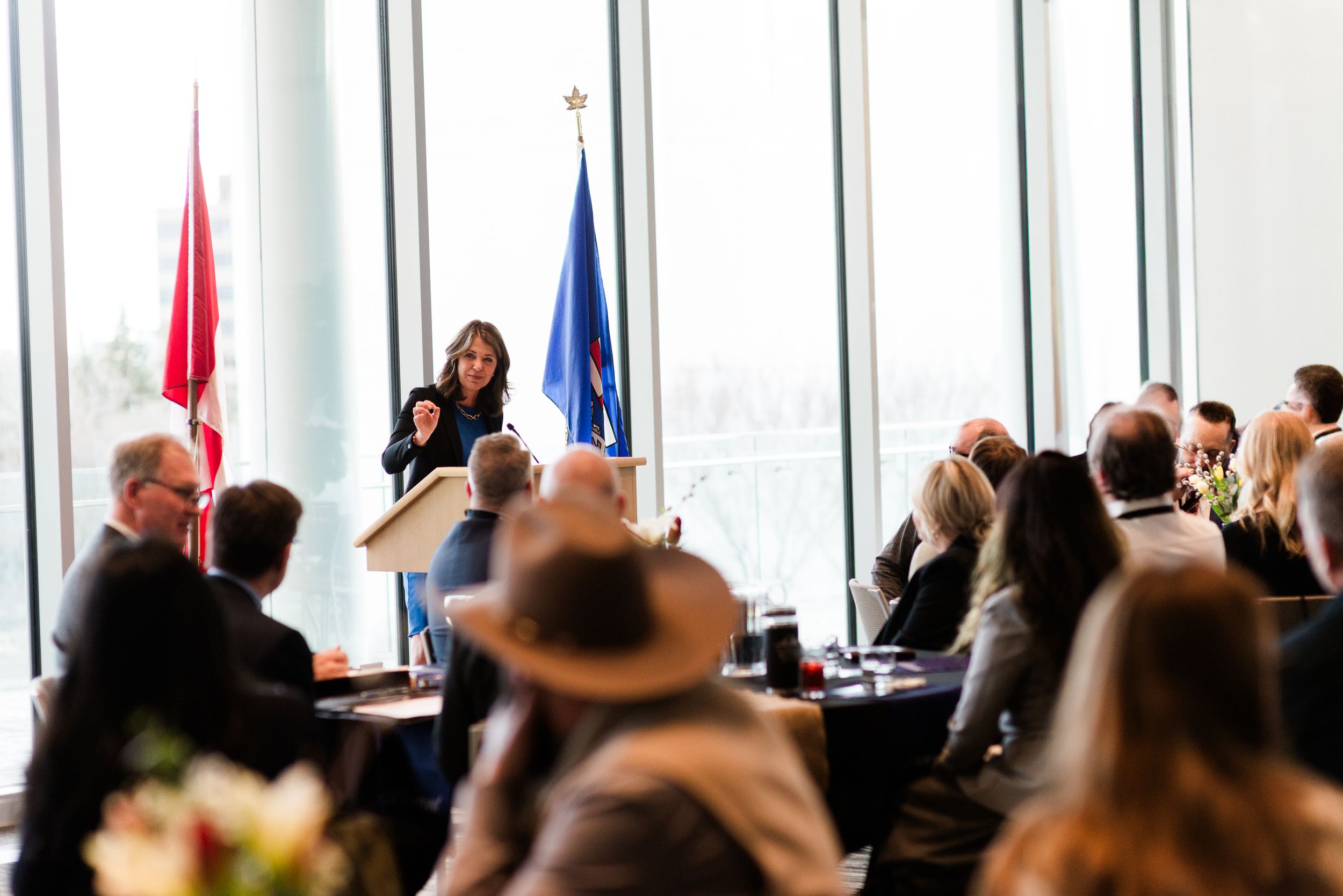At a formal meeting or conference, attendees gather before flags while someone speaks at a podium to the audience.