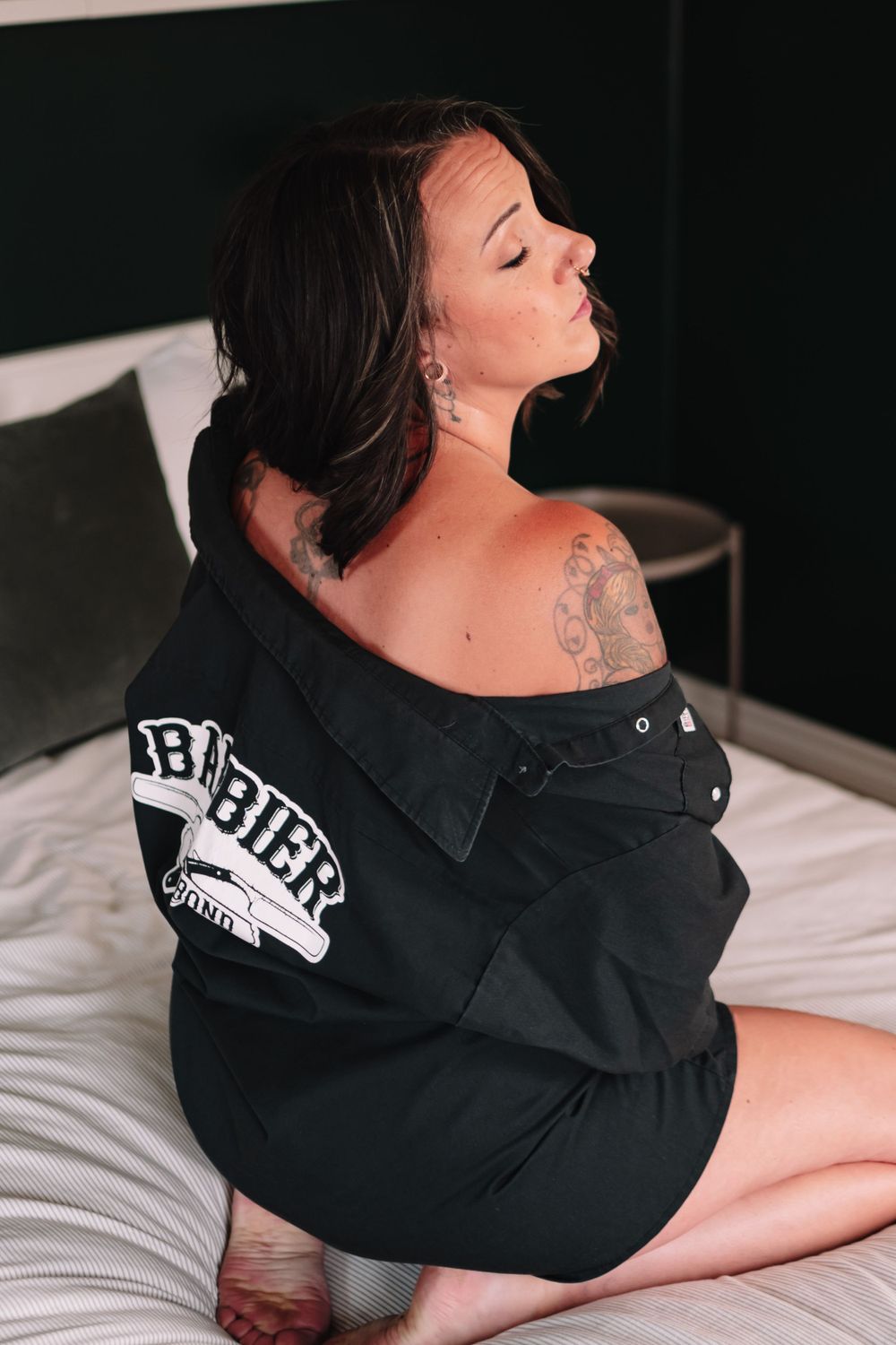 Side view of someone in dark oversized hoodie posing on white bedding in moody boudoir photo.