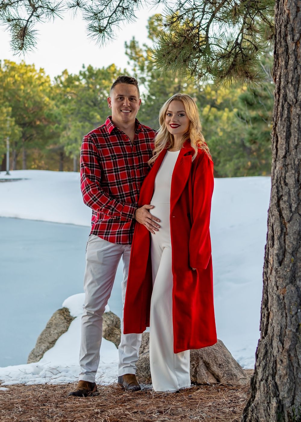 A couple poses by pine trees in winter, with one wearing a bright red coat against snowy ground.