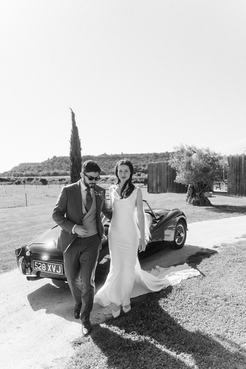 A romantic black and white wedding photo series showing a couple posing playfully by a vintage car in a rural setting.