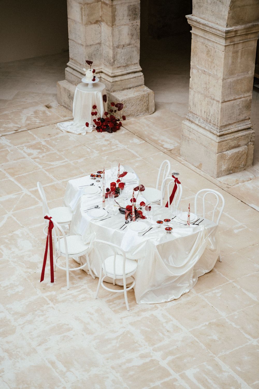 Elegant white wedding table setting with red floral accents and ribbons in a stone pillared venue.