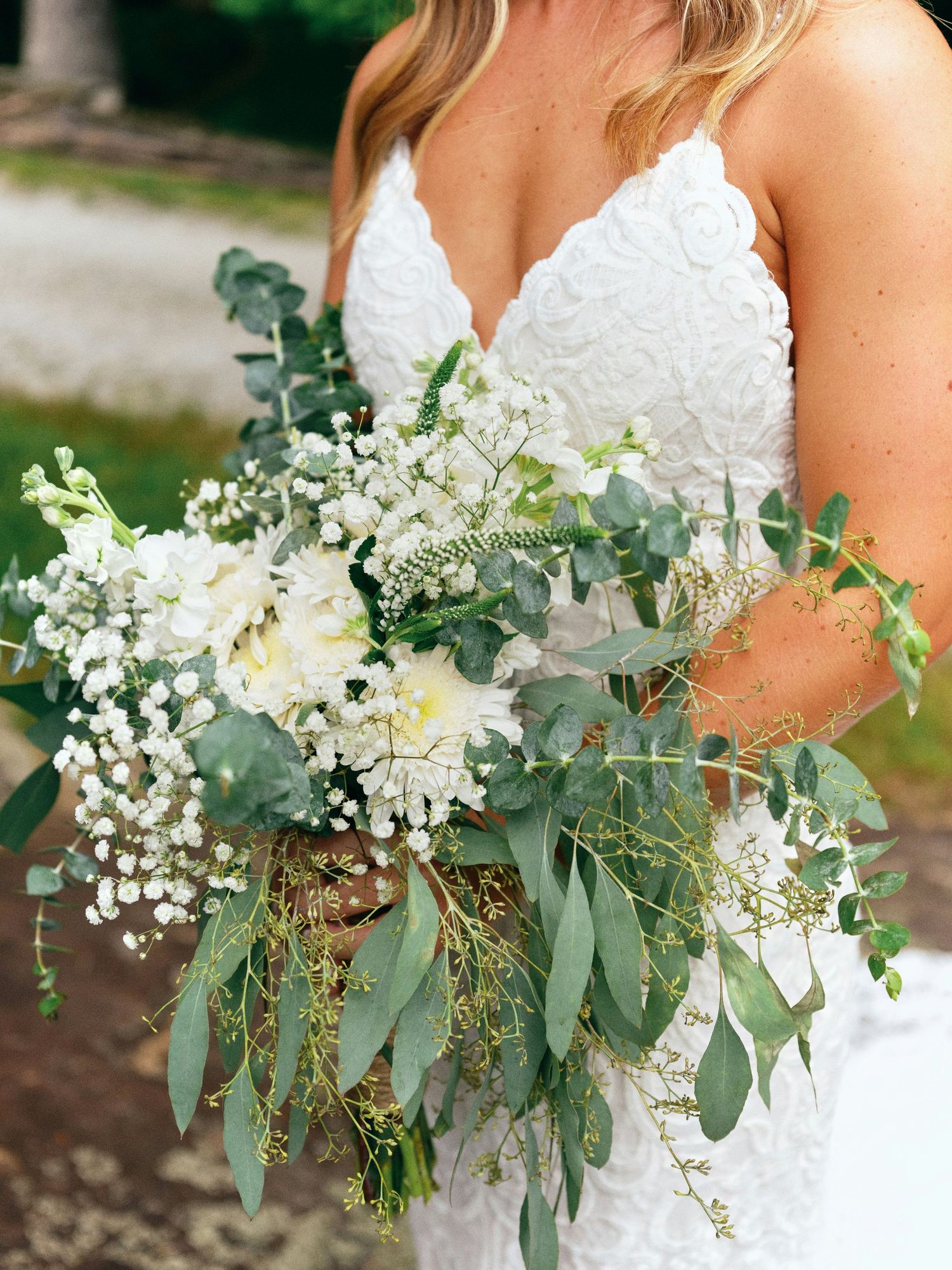A lush bridal bouquet featuring white flowers, baby's breath and cascading eucalyptus leaves against a white lace dress.