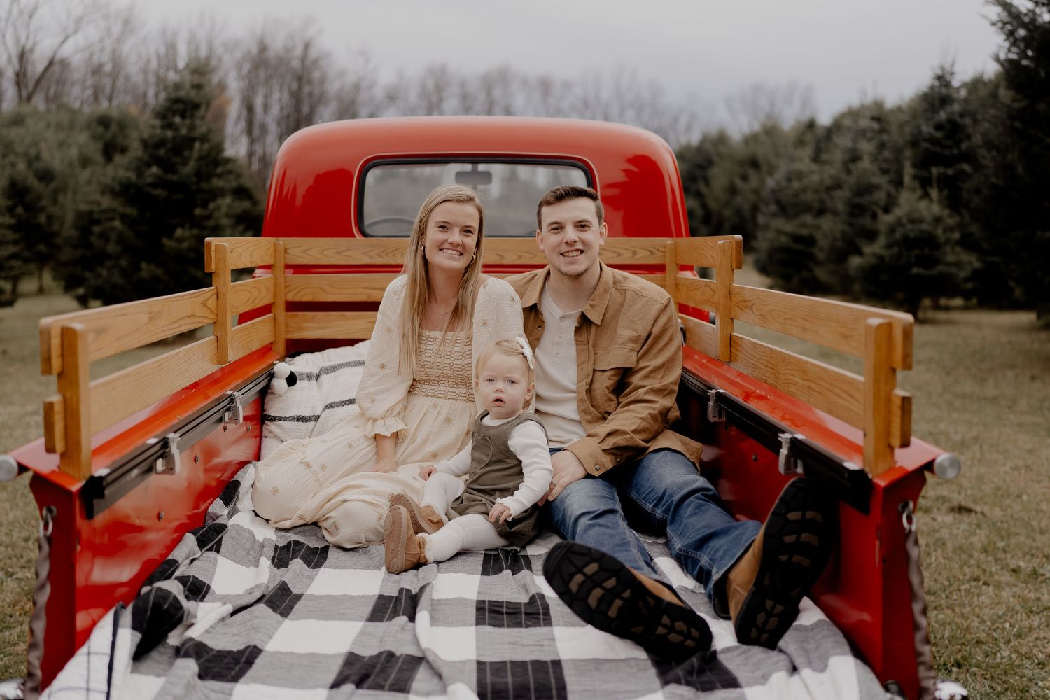 A couple sits on a plaid blanket in the back of a vintage red pickup truck in a rural outdoor setting.