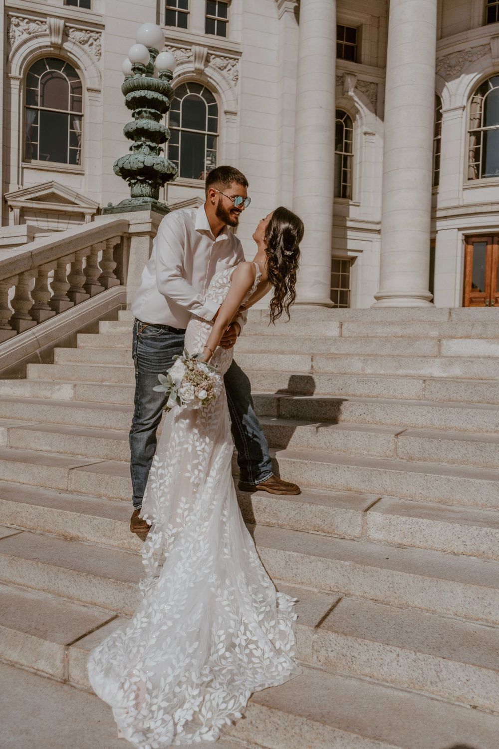 Groom dipping bride in a wedding gown on sunlit steps of a grand historic building.