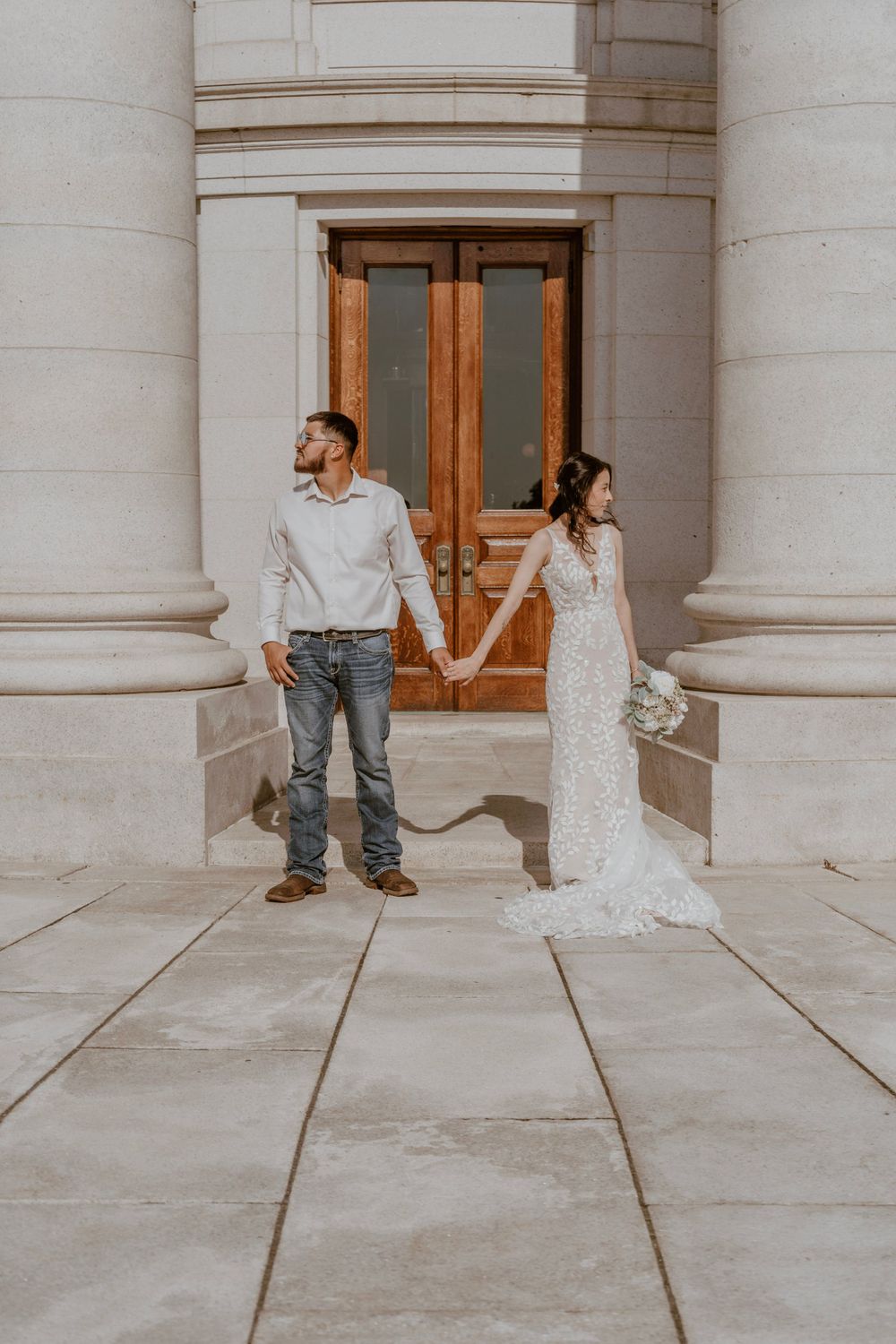 Bride and groom hold hands standing apart before grand wooden doors and stone columns.