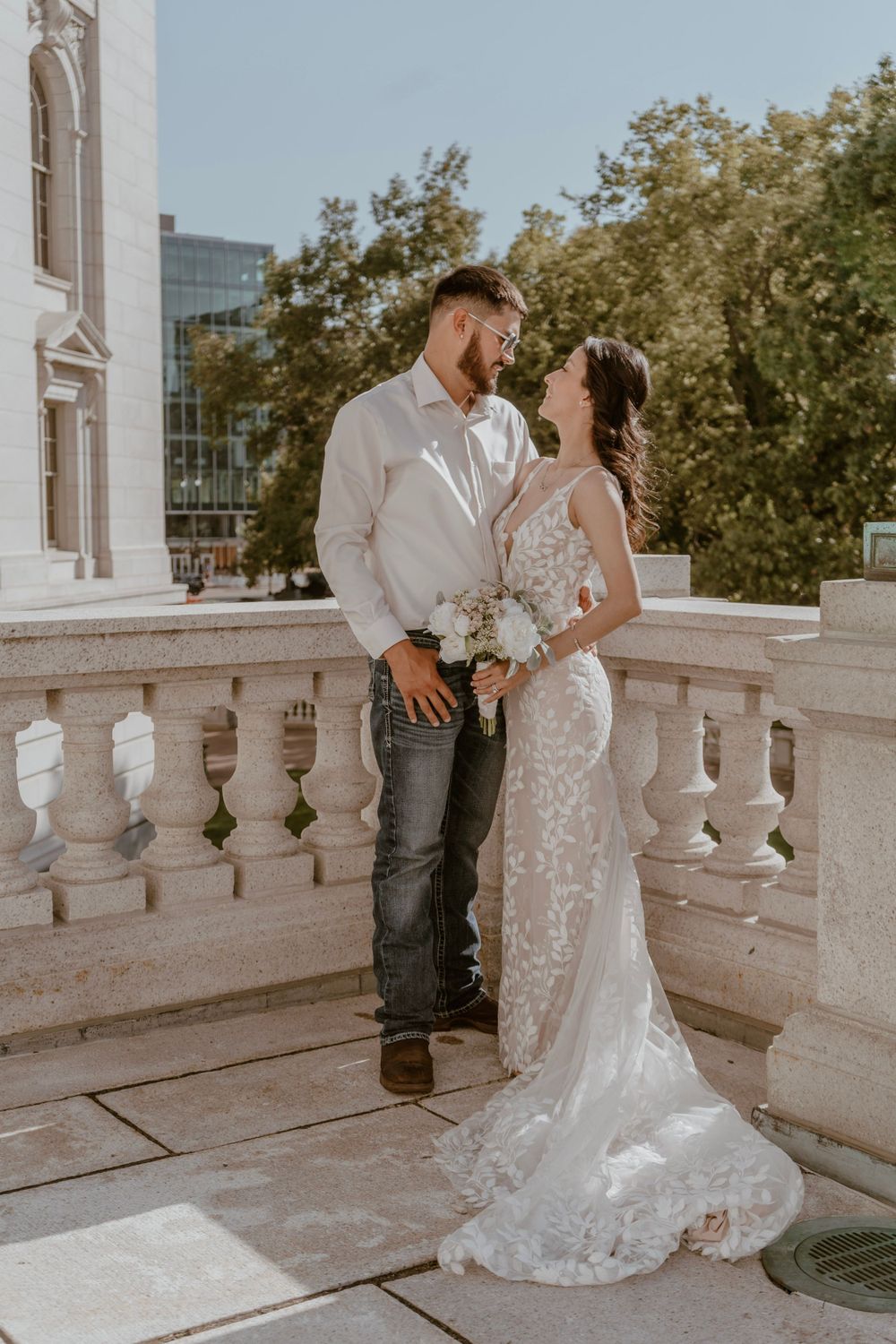 Bride and groom share a romantic gaze on a sunlit stone balcony with lush trees.