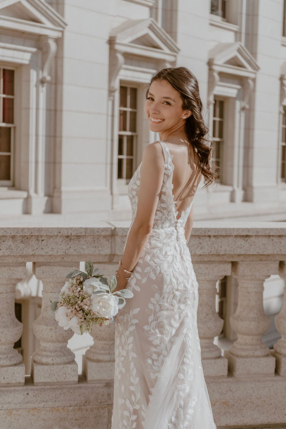 Smiling bride in lace gown holds white bouquet on stone balcony of elegant building.