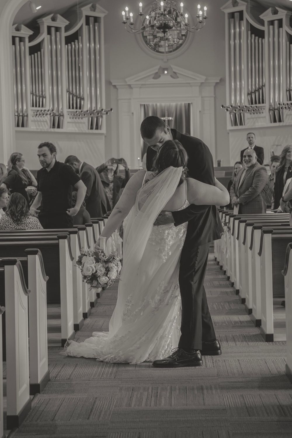 Black and white photo of newlyweds sharing their first kiss in a beautiful traditional church aisle.