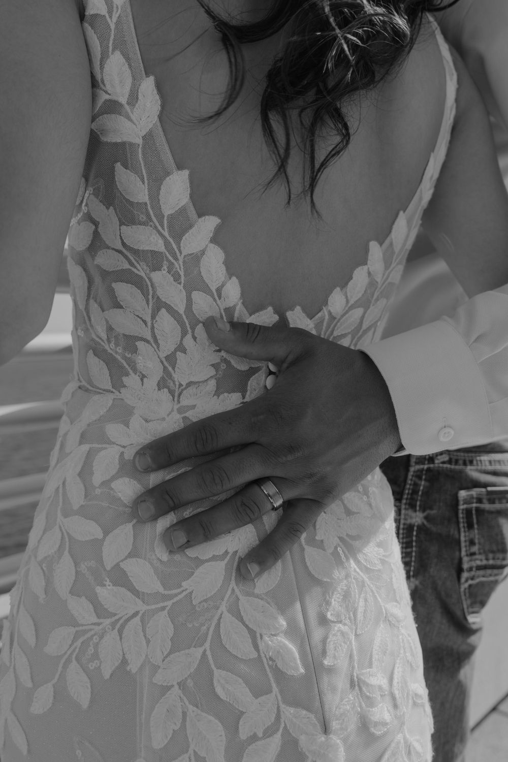Black and white close-up of groom's hand with wedding ring resting on bride's lace gown.