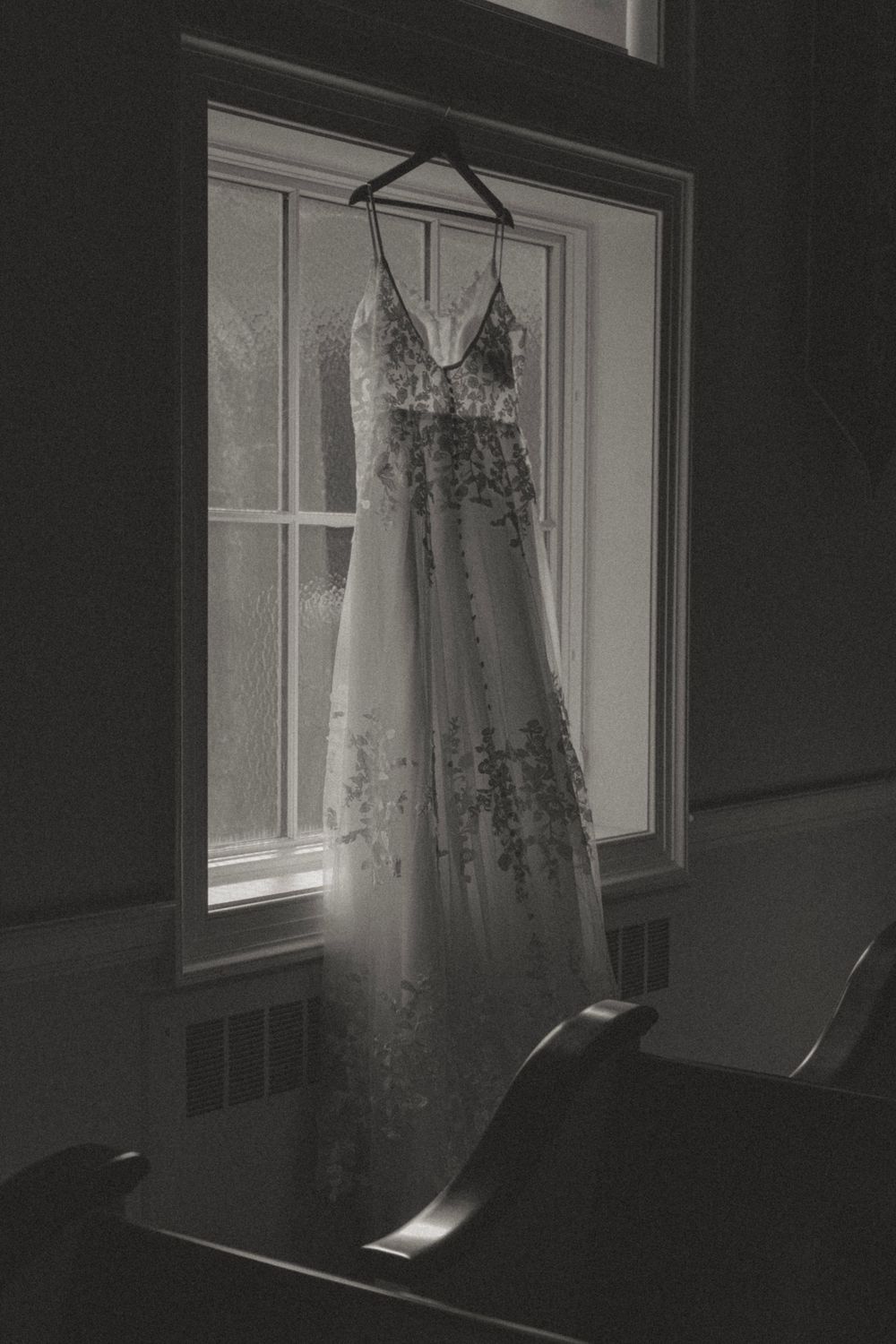Black and white photo of an elegant lace wedding gown hanging in a sunlit church window.
