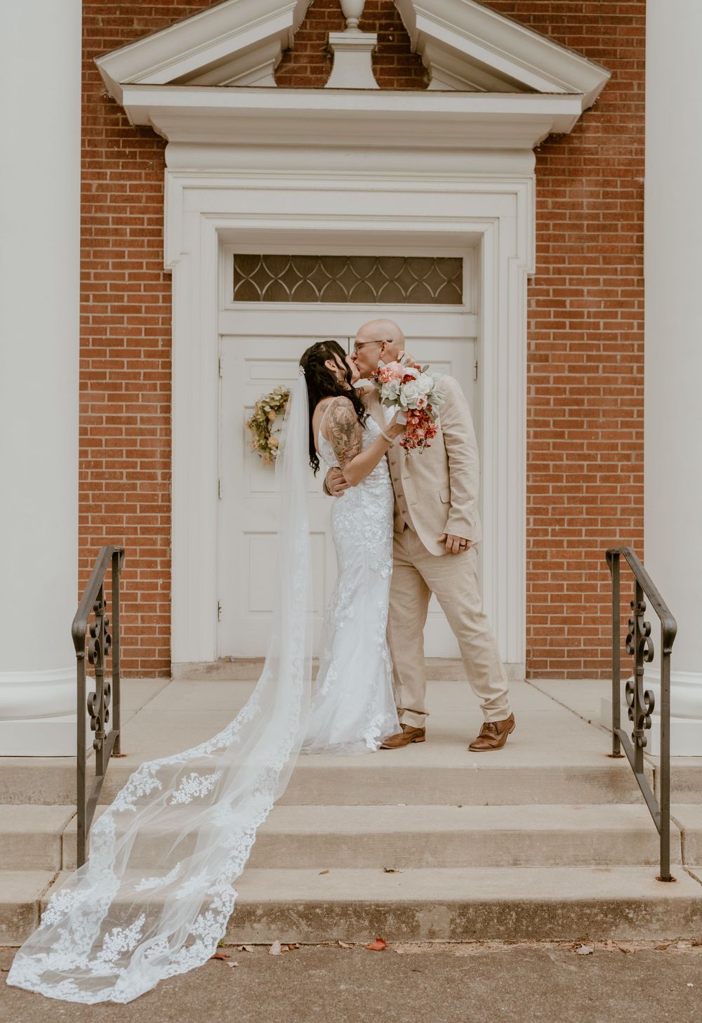 Bride and groom kiss on church steps, her long lace cathedral veil trailing behind.