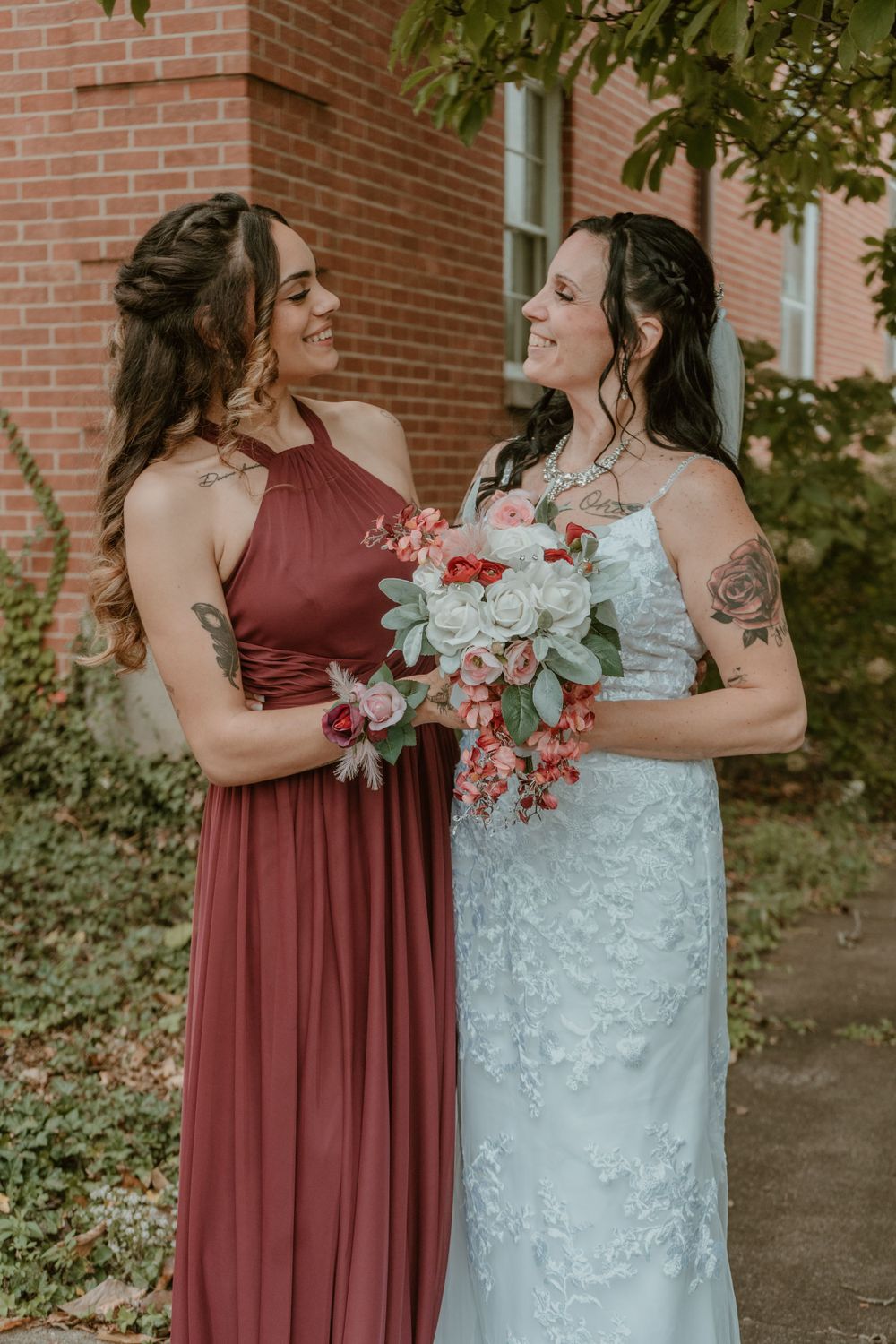 Bride in white lace gown and bridesmaid in burgundy dress smile warmly at each other.