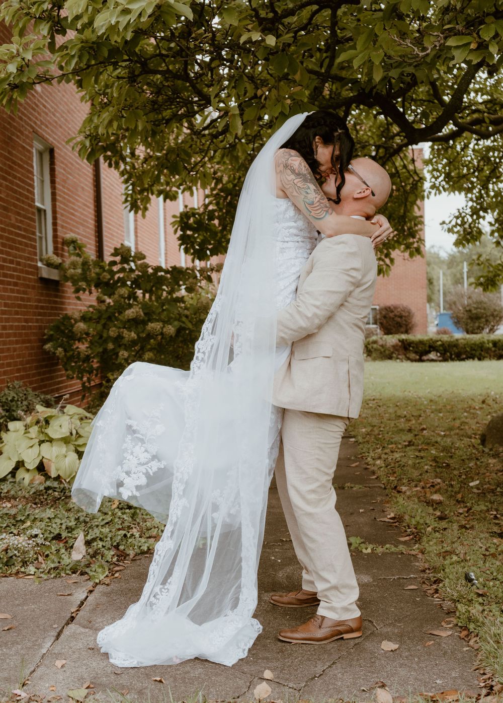 Groom lifts bride in lace gown and veil outdoors near brick building and green trees.