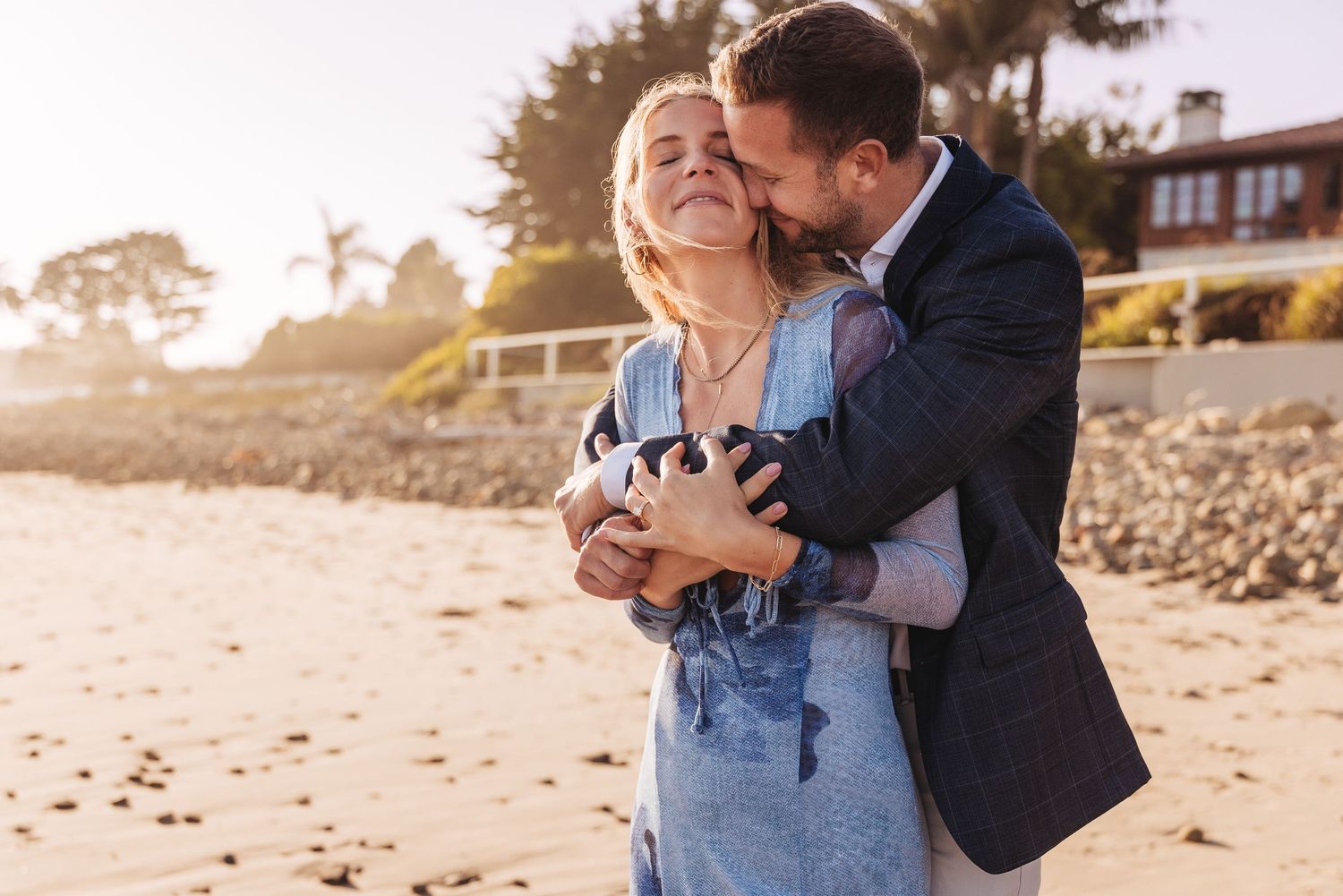 Romantic couple embraces on beach with two horses in background near coastal cliffs at sunset.