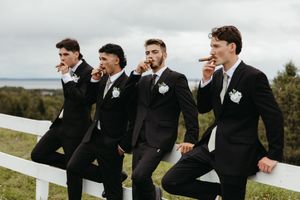 Groom and groomsmen pose on fence overlooking Bay of Fundy smoking cigars fine art wedding photography in New Brunswick, editorial style
