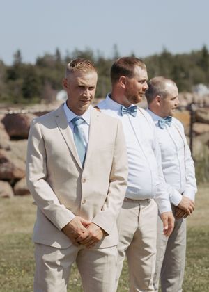Groom and groomsmen stand during wedding ceremony backyard ocean front wedding in Cap Pele New Brunswick, summer full sun wedding