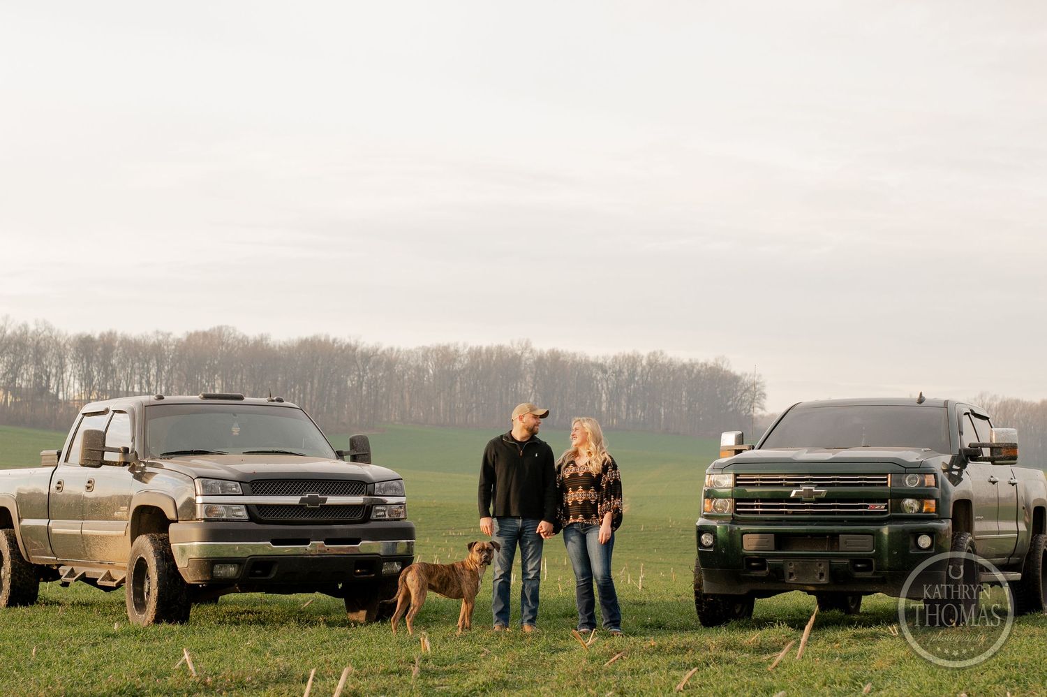 Two couples with their dogs stand between black pickup trucks in an open field.