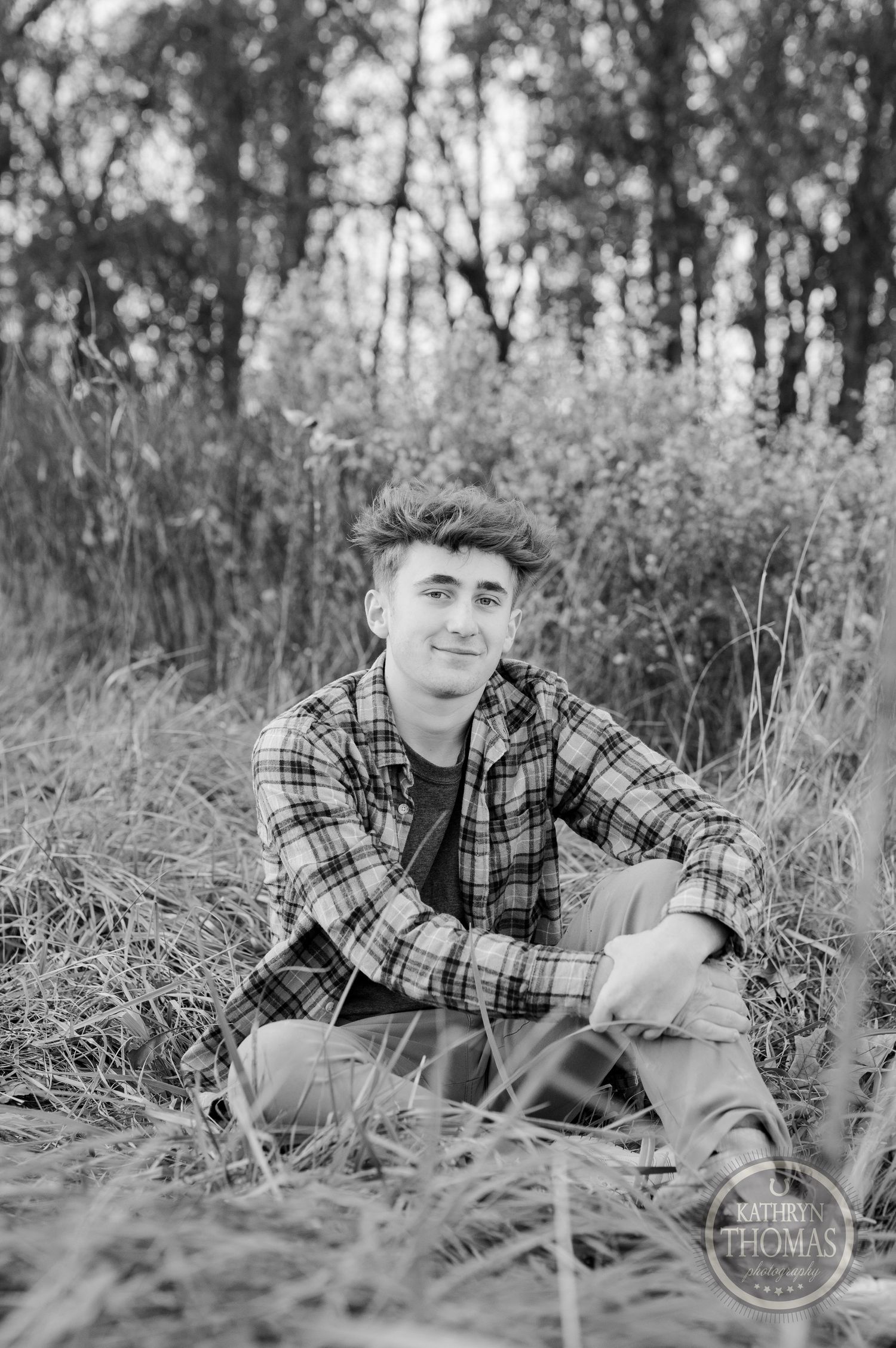A black and white portrait of someone sitting in tall grass wearing a plaid shirt.